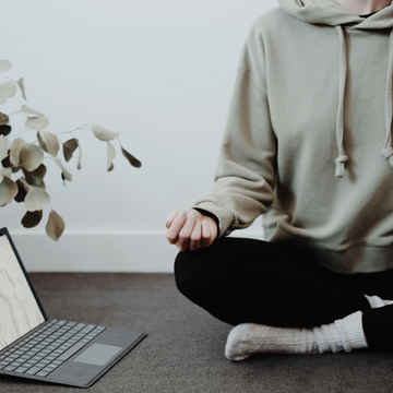 Man in tracksuit meditating cross legged on grey carpet with laptop next to him in his office wellbeing space